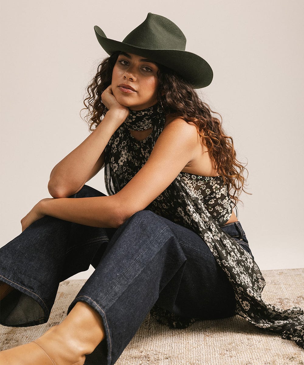 Woman wearing a green cowboy hat, floral scarf, and blue jeans sitting on a textured surface. #color_dark green