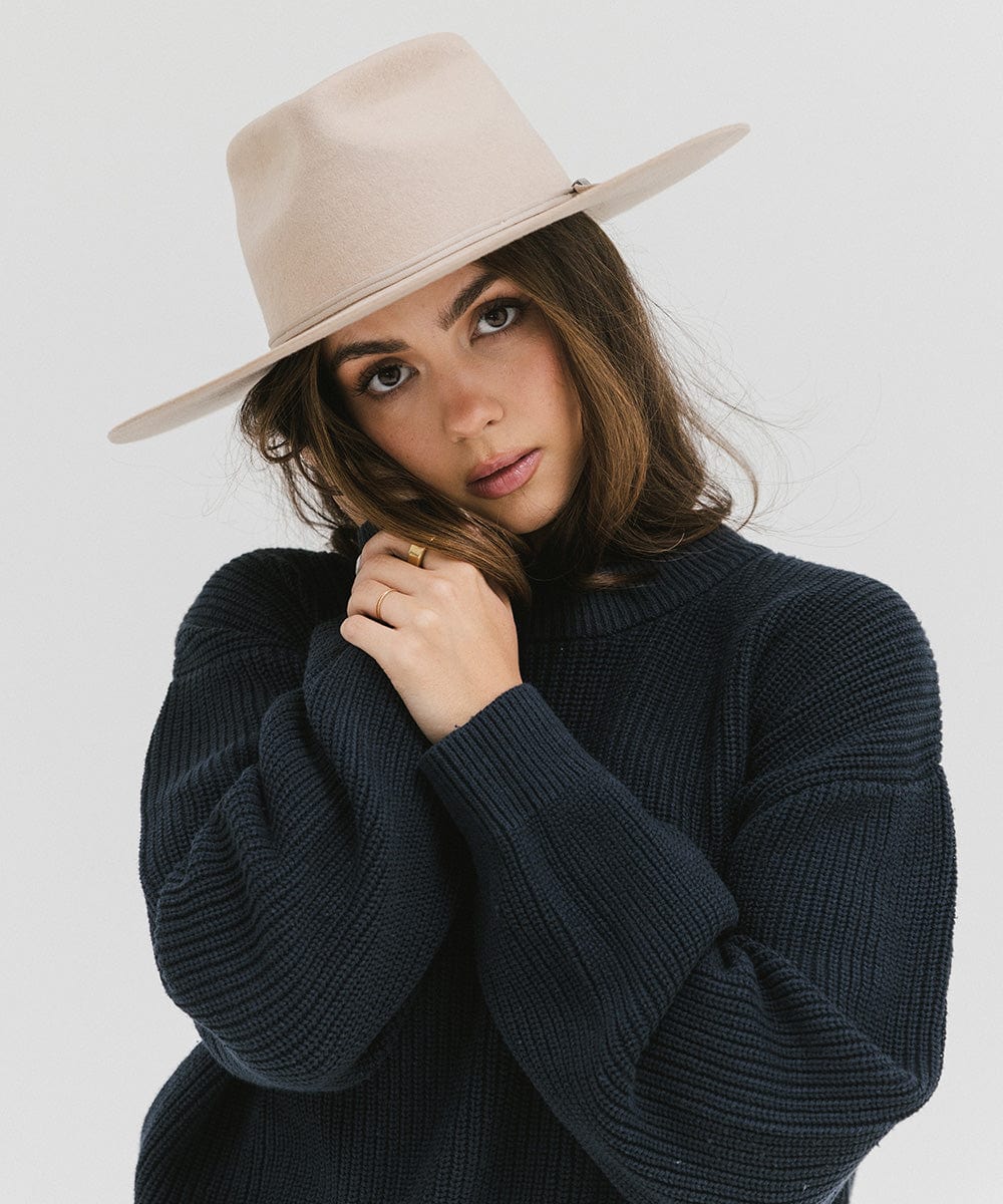 Woman wearing a cream fedora hat with a hat band and a dark blue sweatshirt against a plain background #color_cream