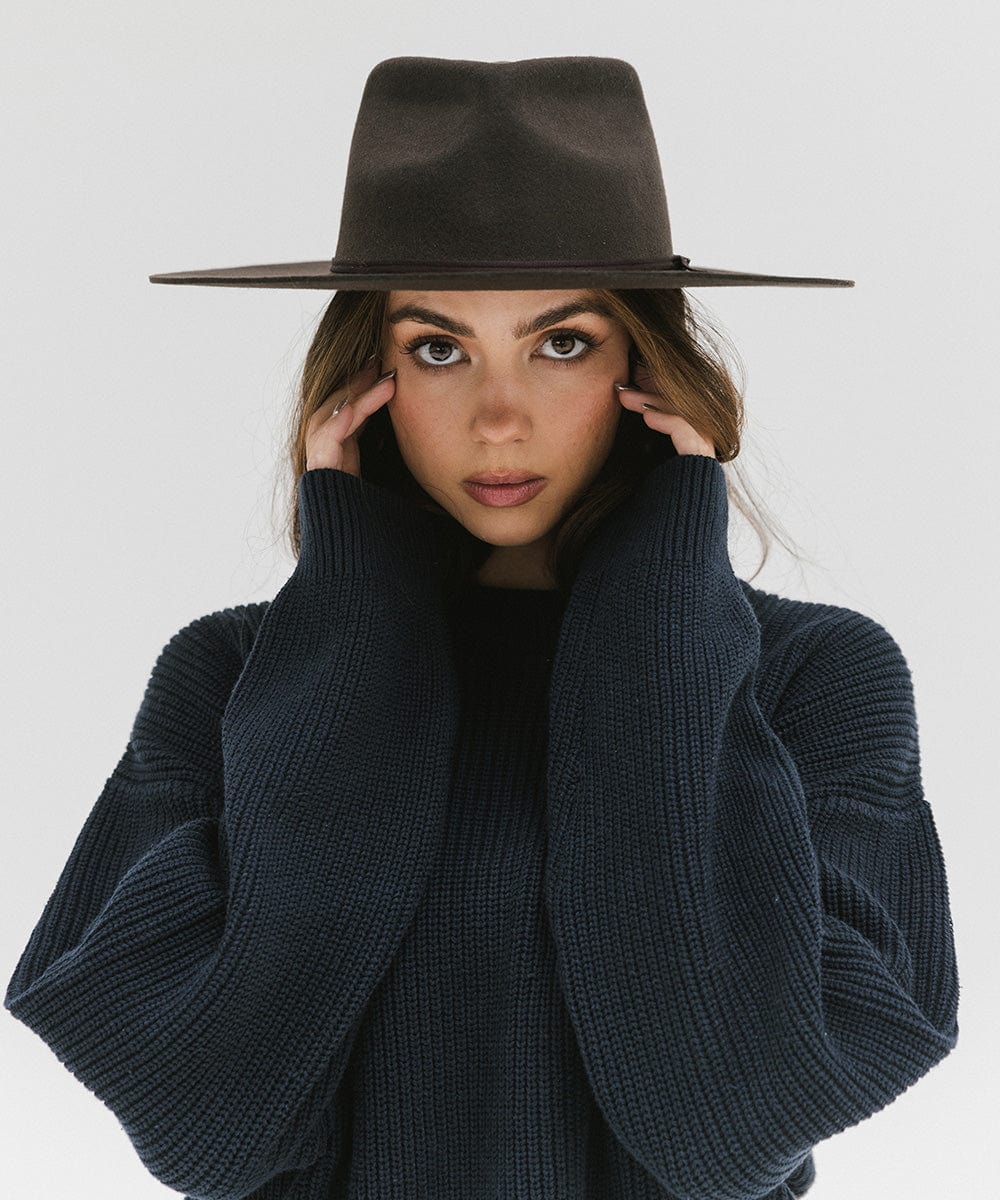 Woman wearing a dark brown fedora hat with a hat band and a dark blue sweatshirt against a plain background #color_dark brown