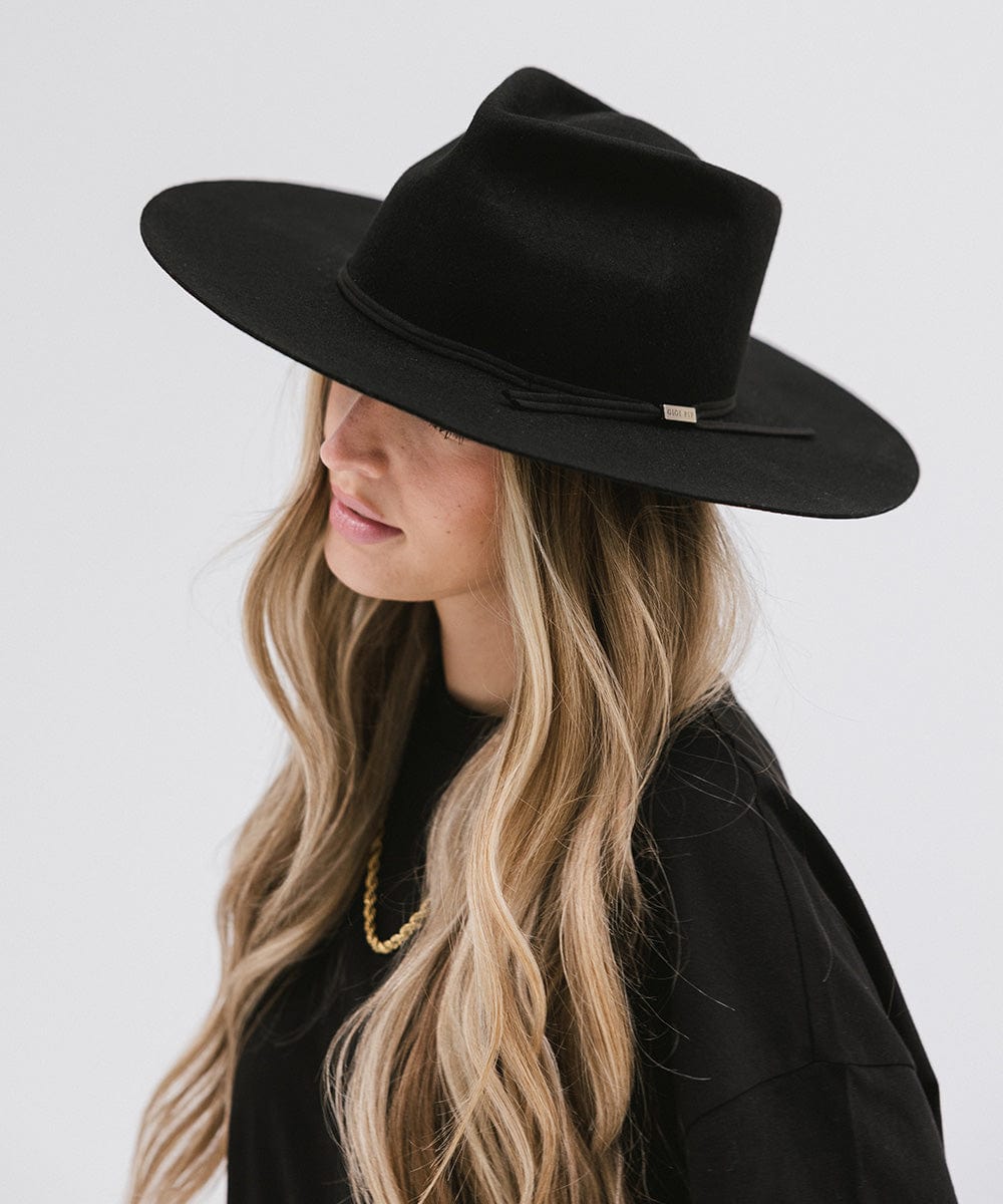 Woman with long hair wearing a black fedora hat with a hat band and a black t-shirt, looking sideways against a plain background #color_black