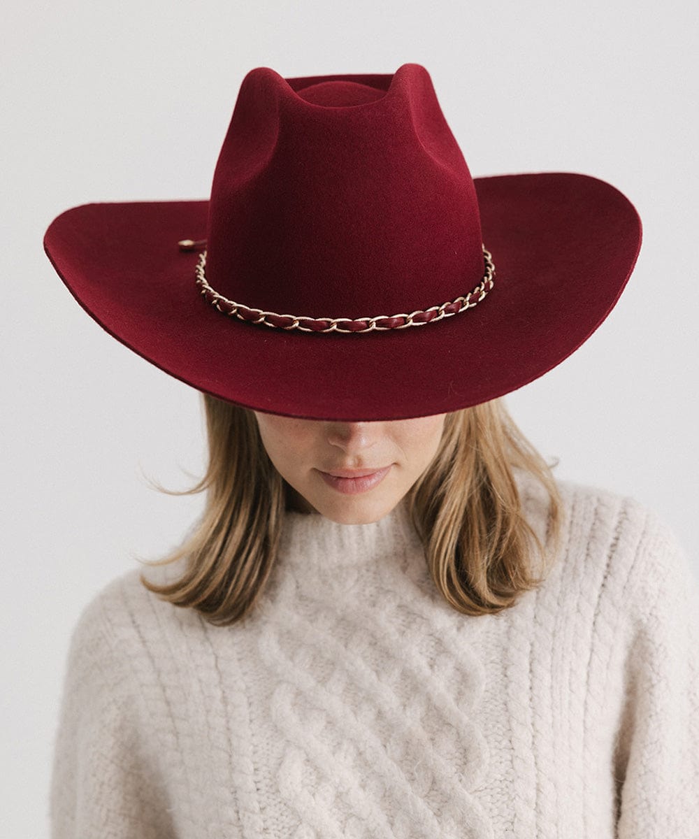 Person wearing a burgundy colored felt hat with a metallic chain detail on the band and a white sweater against a plain background #color_burgundy