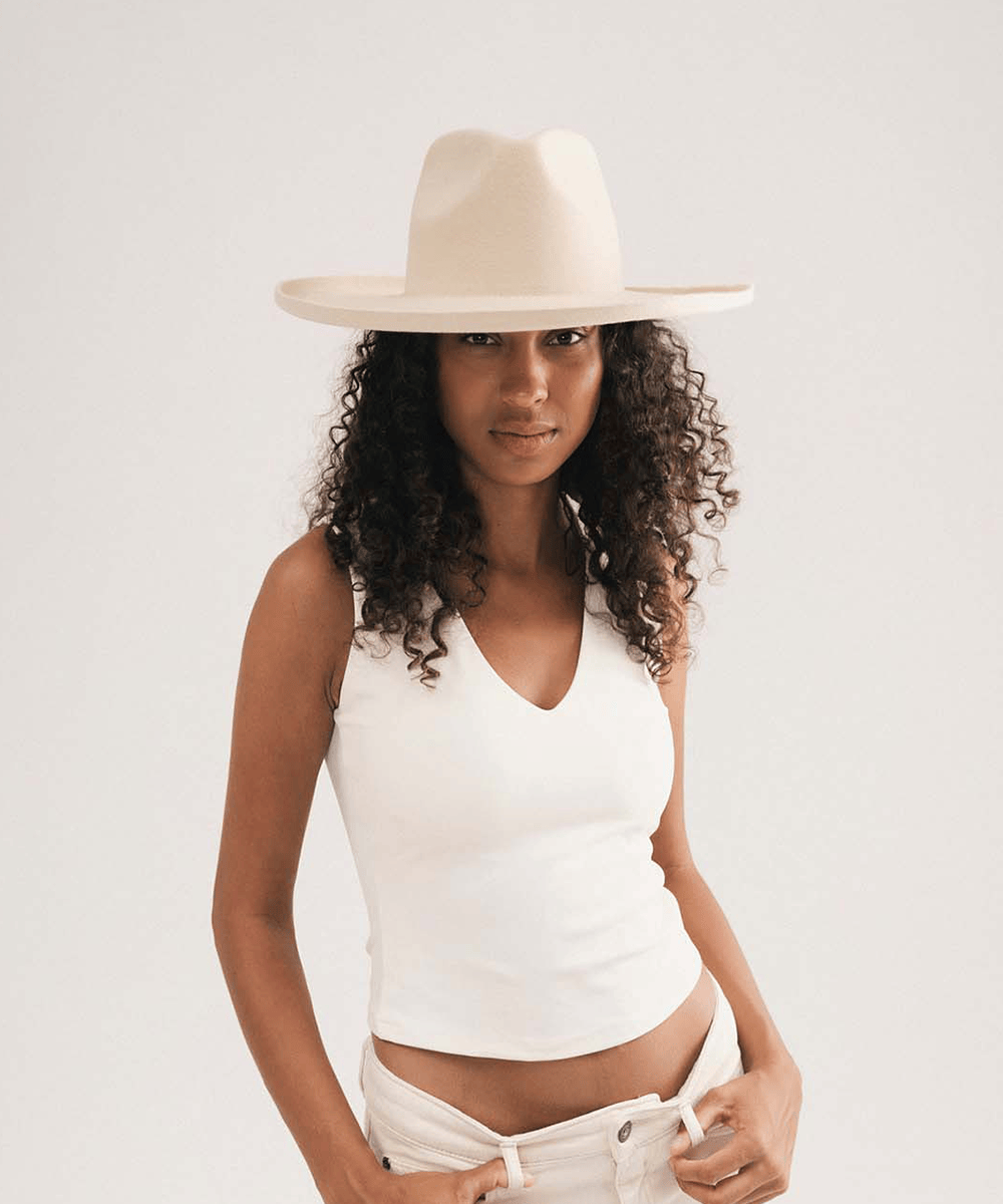 A woman wearing a cream-colored wide-brim fedora hat, a fitted white ribbed tank top, and matching cream pants, posed against a solid light gray studio background #color_cream