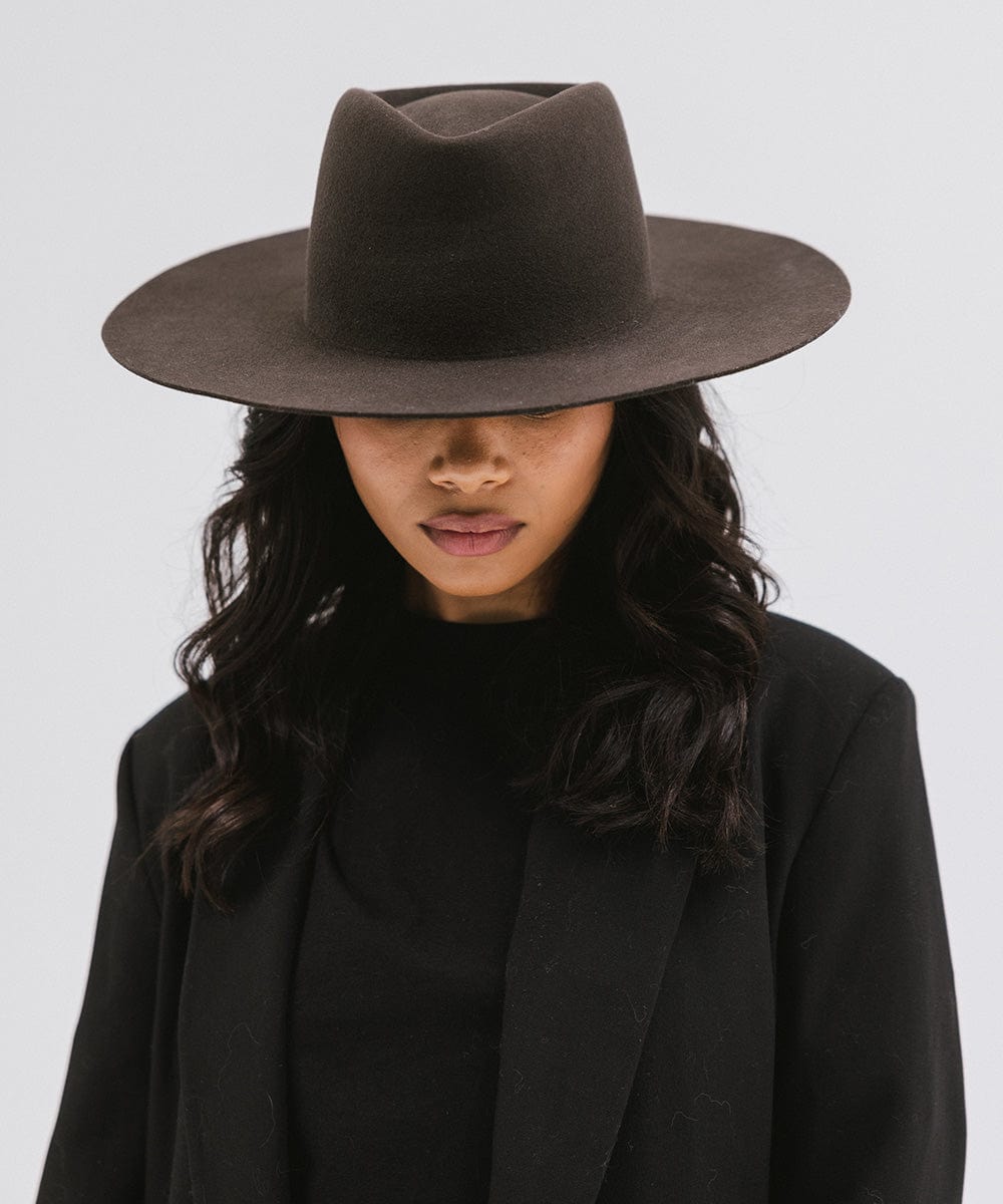 Woman wearing a dark brown wide brim felt hat with a triangle crown and a black t-shirt with a black blazer, looking down against a white background #color_dark brown