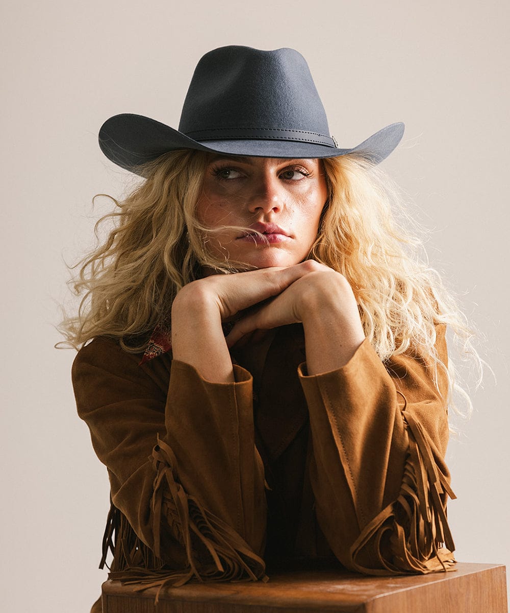 Woman wearing a vintage blue colored cowboy hat and brown jacket, sitting on a wooden block against a plain background #color_vintage blue