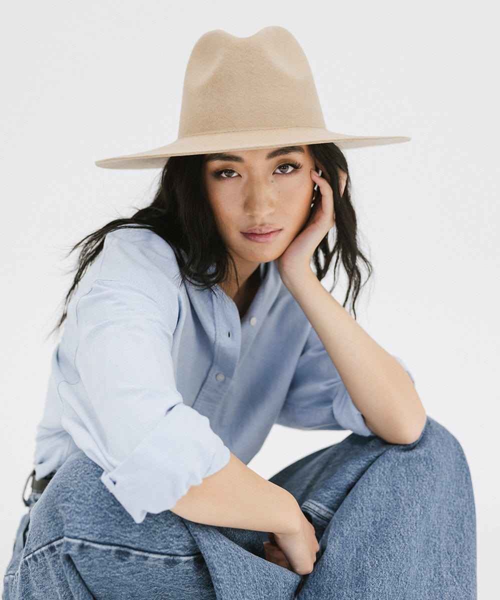 Woman wearing a tan wide brim fedora hat, a blue shirt, and blue jeans against a plain background #color_tan