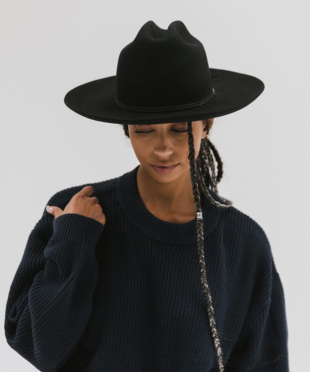 Woman wearing a black Western hat with a classic cattleman crown and a black hat band, and a dark blue sweatshirt, looking down against a plain background #color_black
