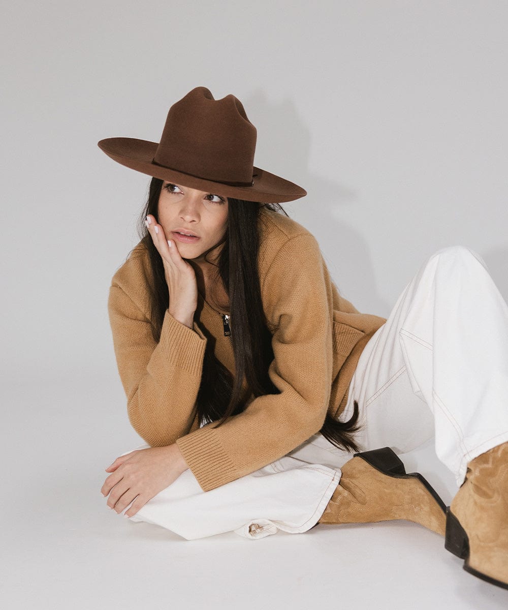 Woman wearing a dark oak colored Western hat with a classic cattleman crown, a brown jacket, white pants and brown boots, sitting against a plain background #color_dark oak
