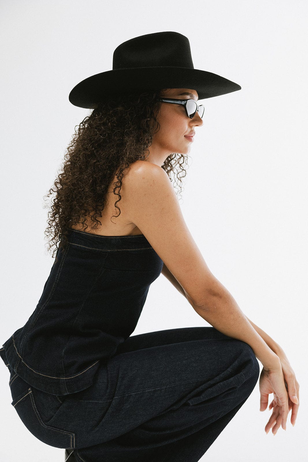 Woman wearing a black cowboy hat, sunglasses, a dark blue denim top and dark blue denim jeans, facing sideways a plain background #color_black