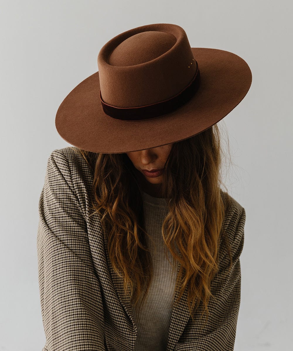 Woman wearing a dark oak colored boater hat with a hat band and a coat, looking down against a plain background #color_dark oak