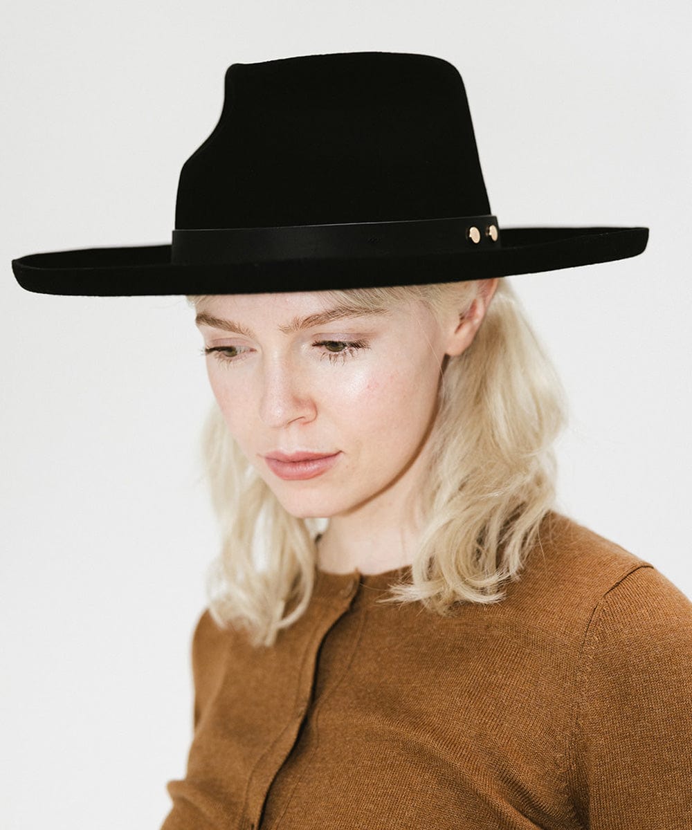 Woman wearing a black felt fedora hat with a hat band and a brown sweater against a plain background #color_black