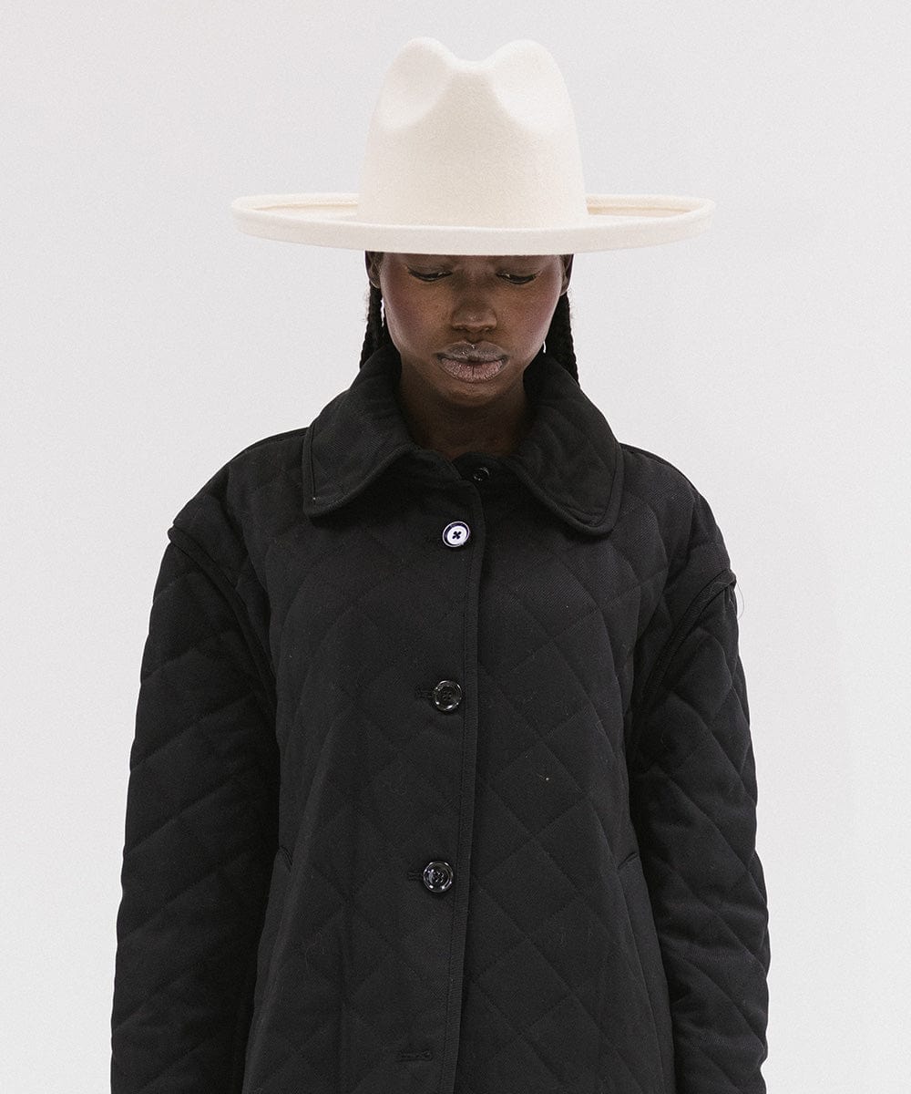 Woman wearing an off white pencil brim felt fedora hat and a black coat against a white background #color_off white