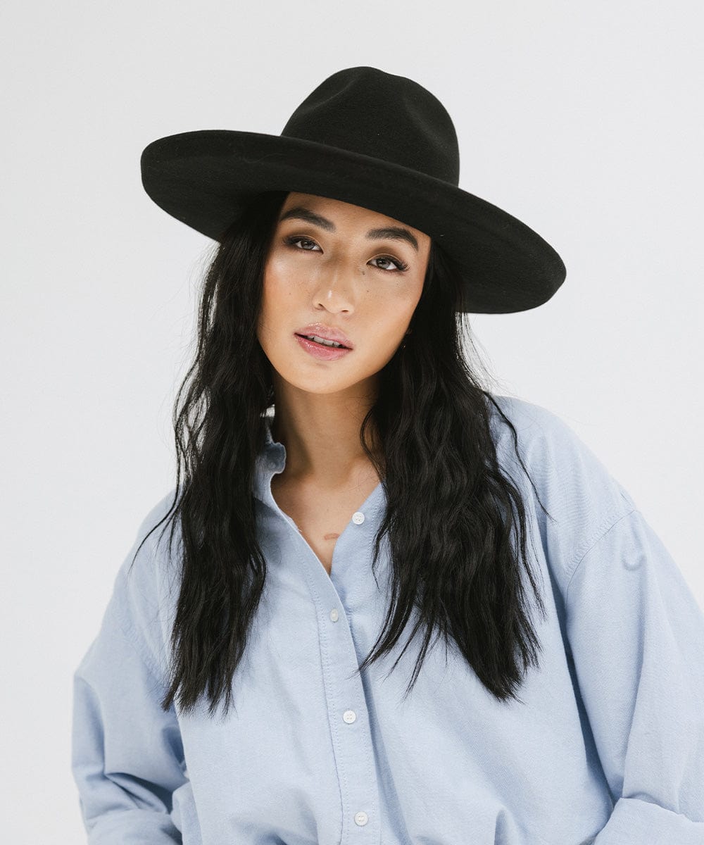 Woman wearing a black pencil brim felt fedora hat and a blue shirt against a white background #color_black