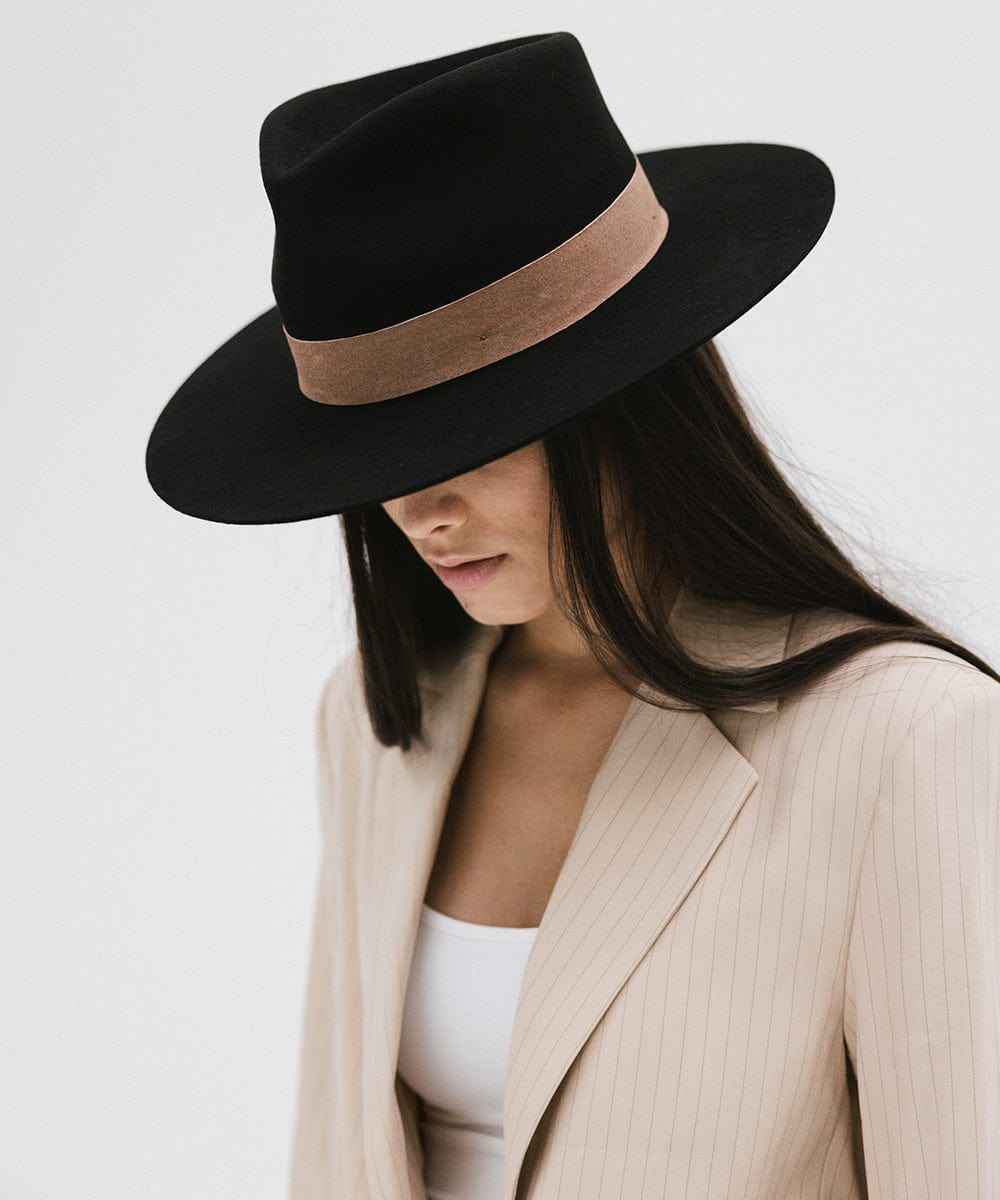 Woman wearing a black fedora hat with a brown band and a cream blazer, looking down against a white background #color_black-brown