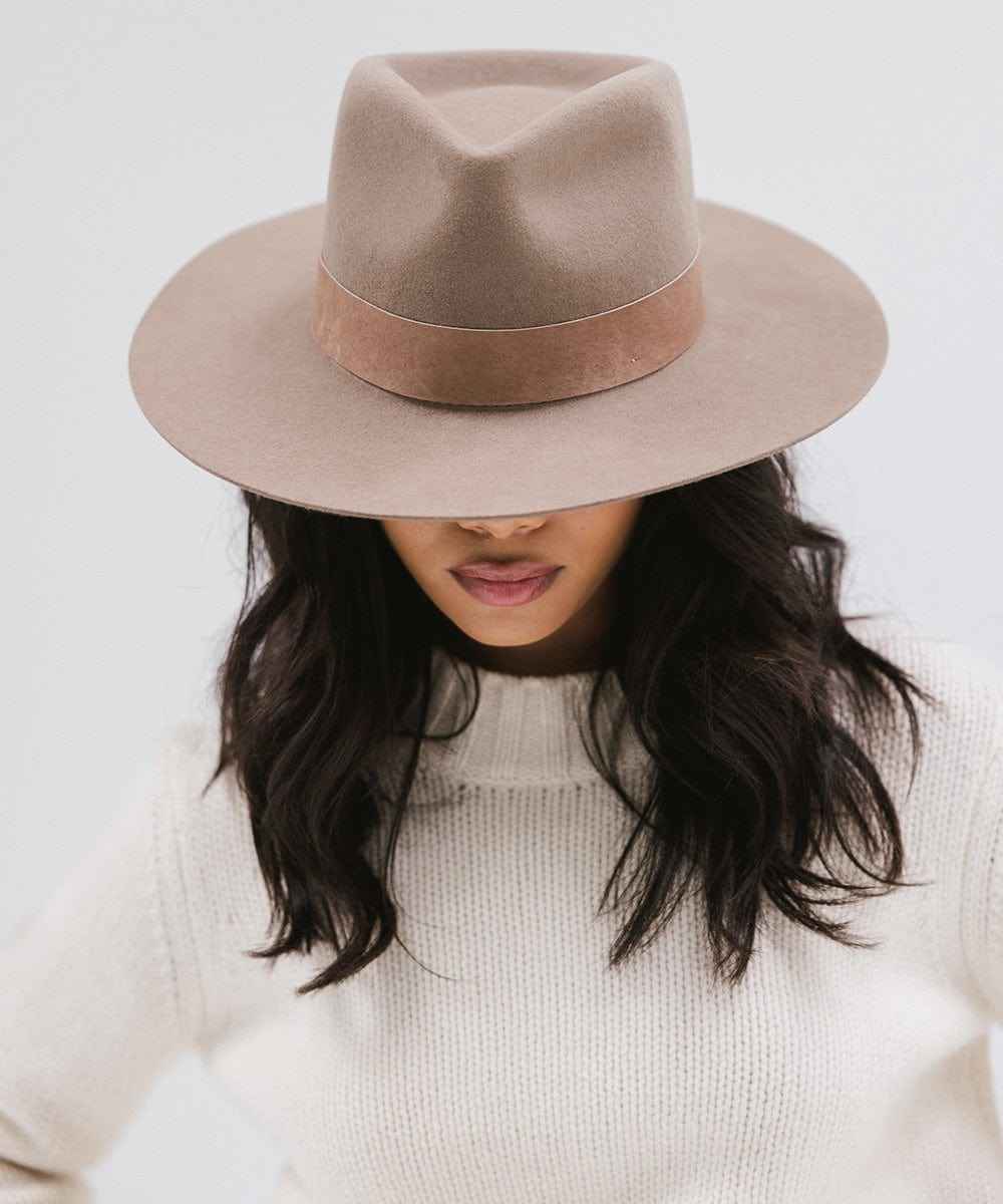 Woman wearing a brown fedora hat with a band and a white sweater, looking down against a plain background #color_brown