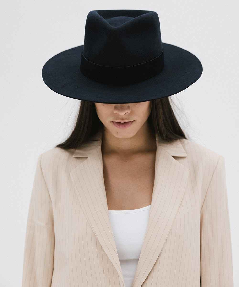 Woman wearing a navy fedora hat with a band and beige blazer, looking down against a white background #color_navy