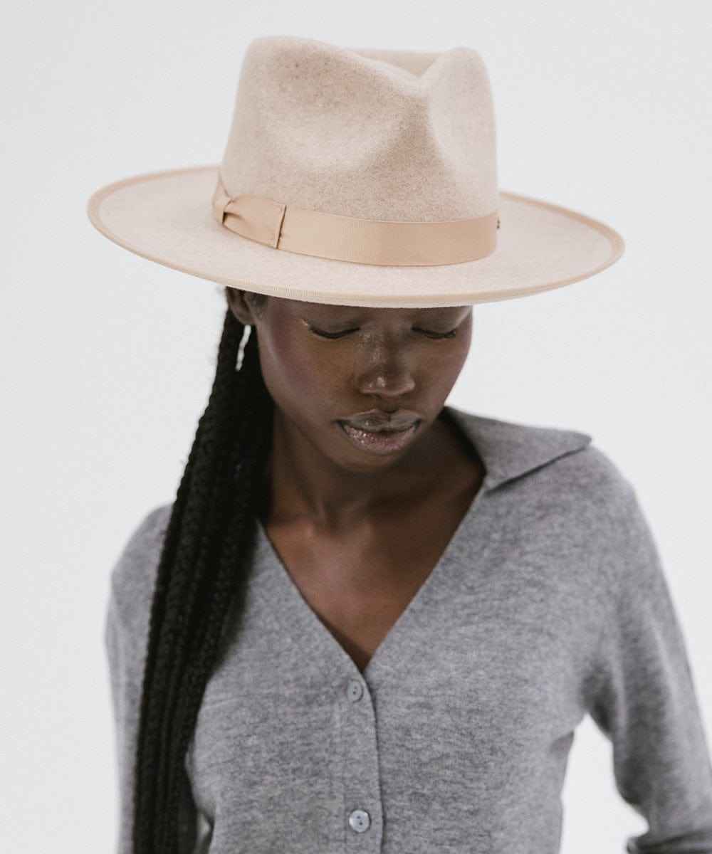 Woman wearing an oatmeal colored fedora hat with a band and a grey shirt, looking down against a plain background #color_oatmeal