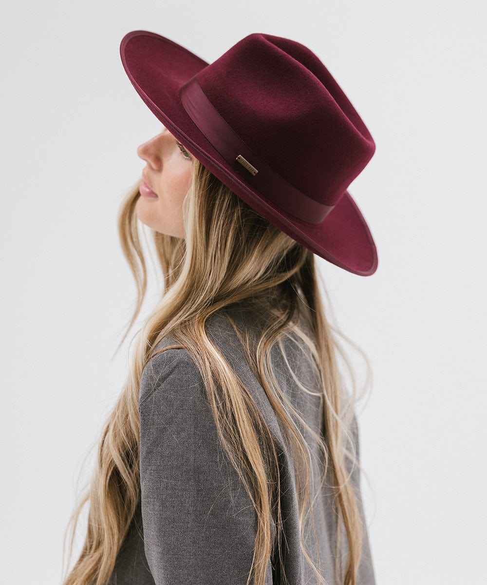 Woman wearing a wine colored fedora hat with a band and a grey blazer, facing sideways against a plain background #color_wine