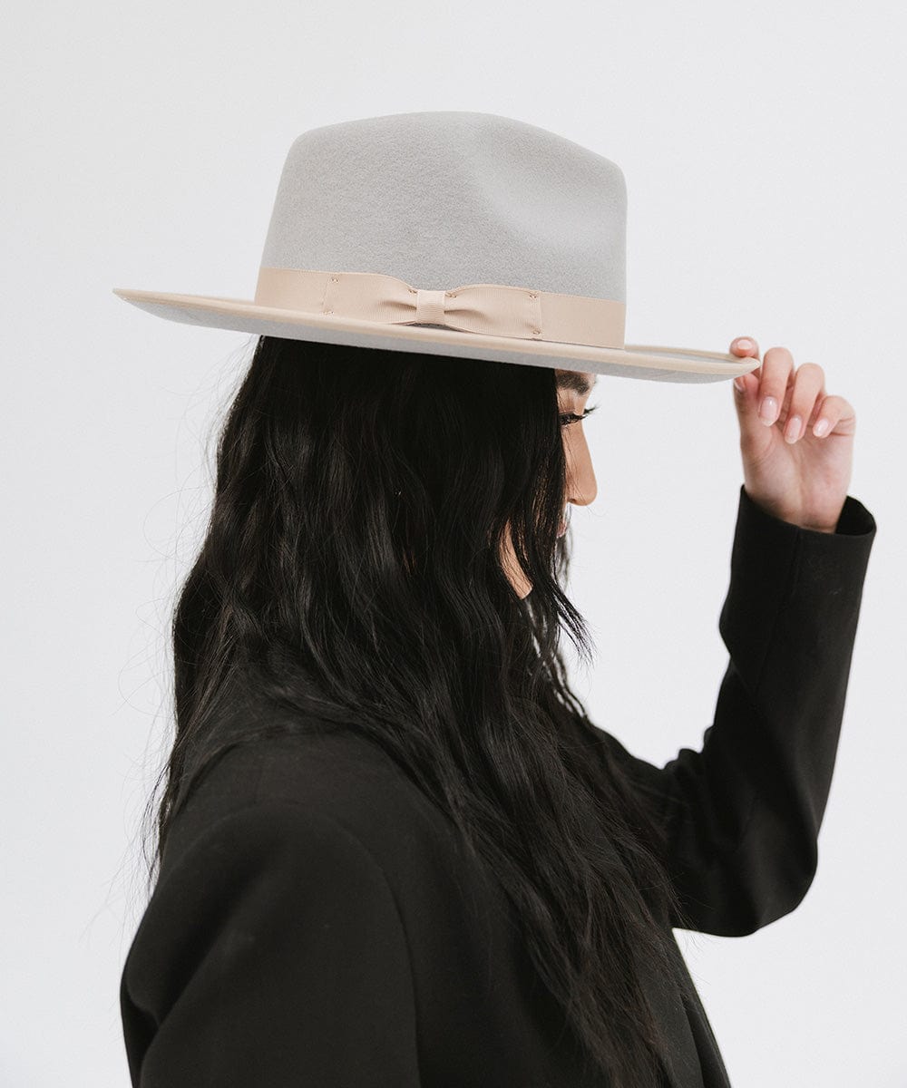 Woman wearing a light grey and tan colored fedora hat with a tan band and a black blazer, facing sideways against a plain background #color_light grey-tan