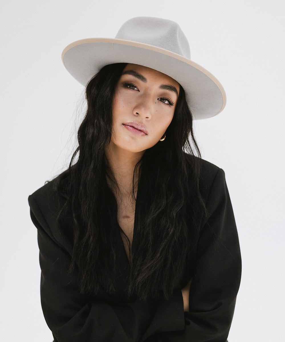 Woman wearing a light grey and tan colored fedora hat and a black blazer against a plain background #color_light grey-tan