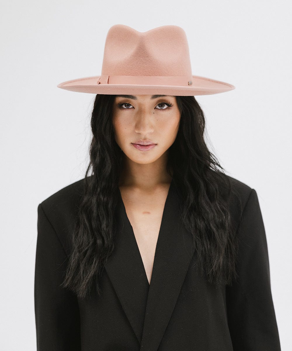 Woman wearing a dusty pink colored fedora hat with a band and a black blazer against a plain background #color_dusty pink