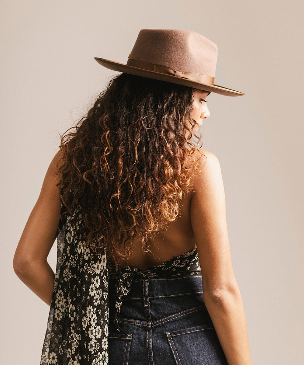 Woman wearing a chocolate colored hat, floral top, and dark blue jeans, facing behind against a plain background #color_chocolate