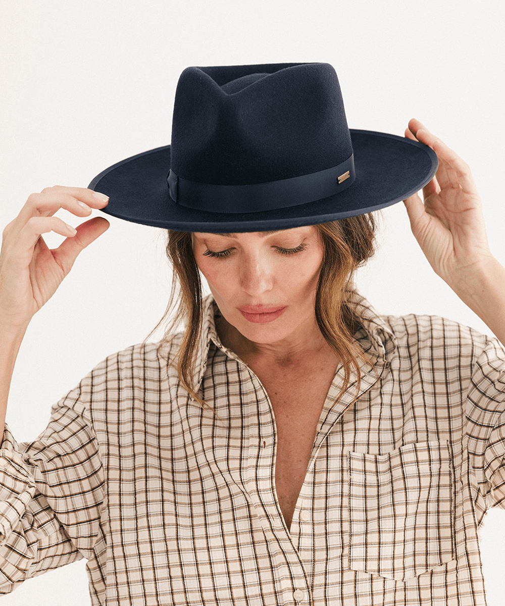 A woman wearing a dark navy blue felt fedora hat with a matching band and a small gold logo tag, holding the hat in place with both hands. She is wearing a beige and brown checkered button-down shirt against a clean white background #color_navy