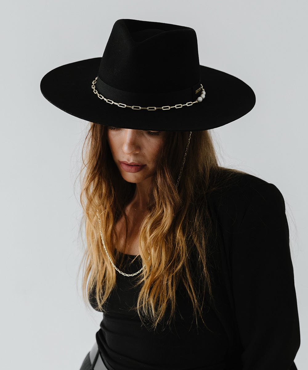 Woman wearing a black wide brim fedora hat with a gold chain detail around the base of the crown and a black top against a plain background #color_black