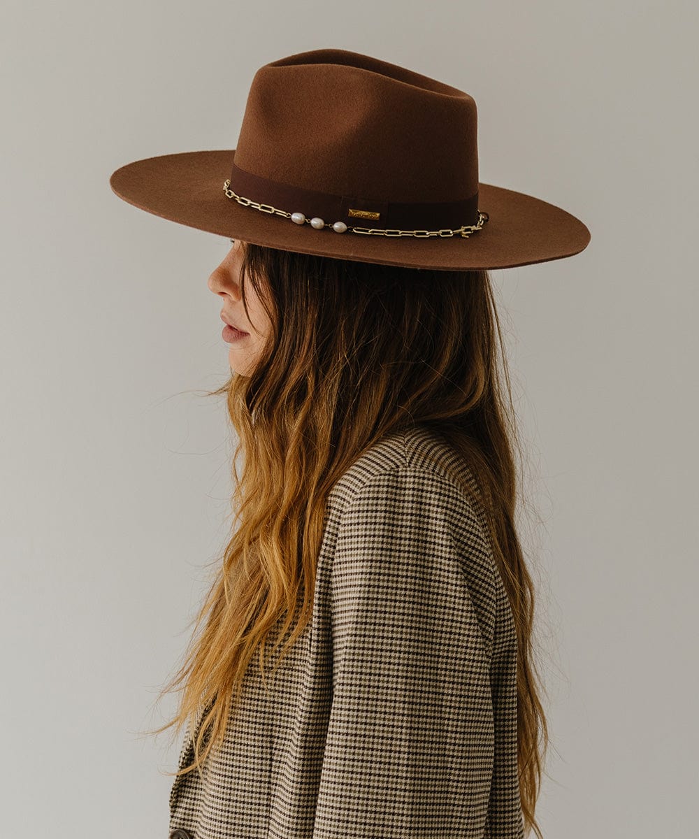 Woman wearing a dark oak colored wide brim fedora hat with a gold chain detail around the base of the crown and a coat, facing sideways against a plain background #color_dark oak