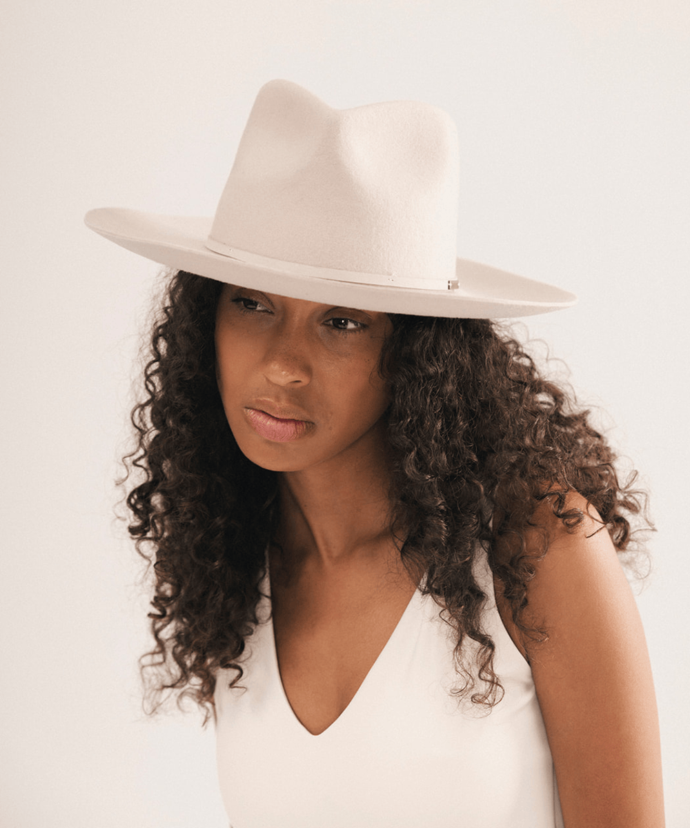 A woman with long, curly dark brown hair wearing a light beige wide-brim fedora hat and a matching cream-colored sleeveless top, photographed against a plain light background #color_ivory