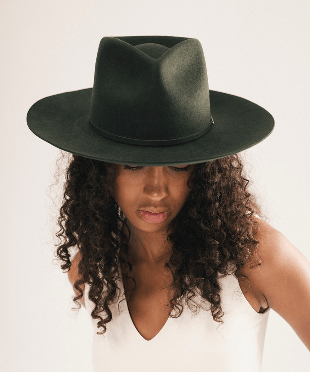 A woman with long, curly dark brown hair wearing a dark forest green wide-brim fedora hat and a white sleeveless top, photographed against a plain light beige background #color_dark green
