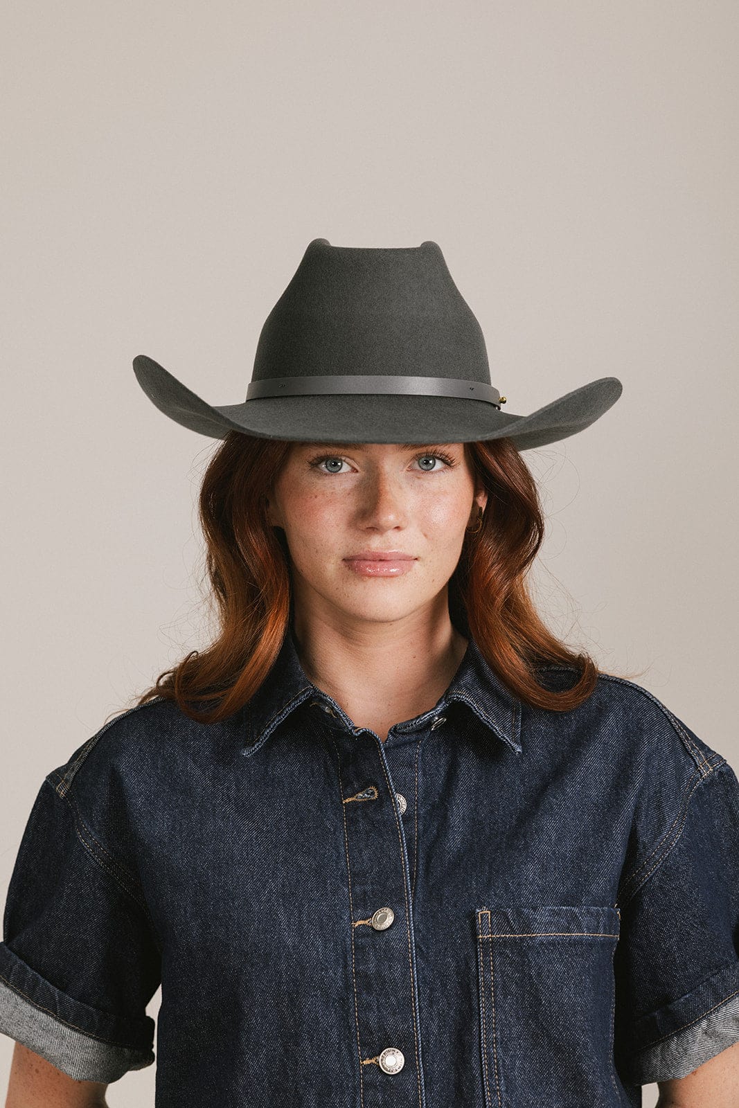 Woman wearing a dark grey cowboy hat with an upturned brim and an attached leather band and a dark blue denim shirt against a white background #color_dark grey