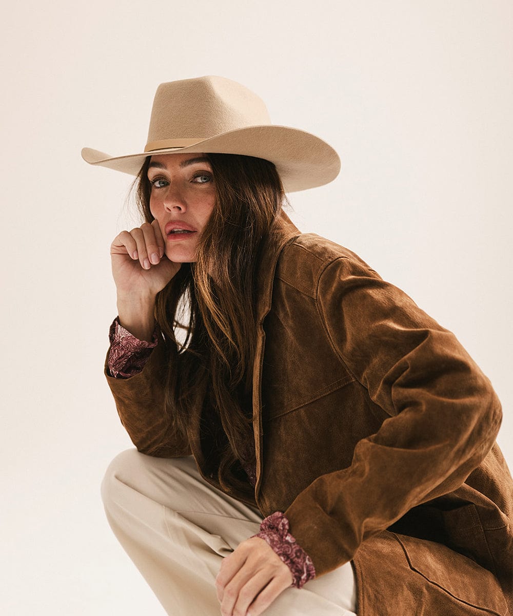 Woman wearing a tan cowboy hat with an upturned brim and an attached leather band, a brown suede jacket and beige pants against a plain background #color_tan