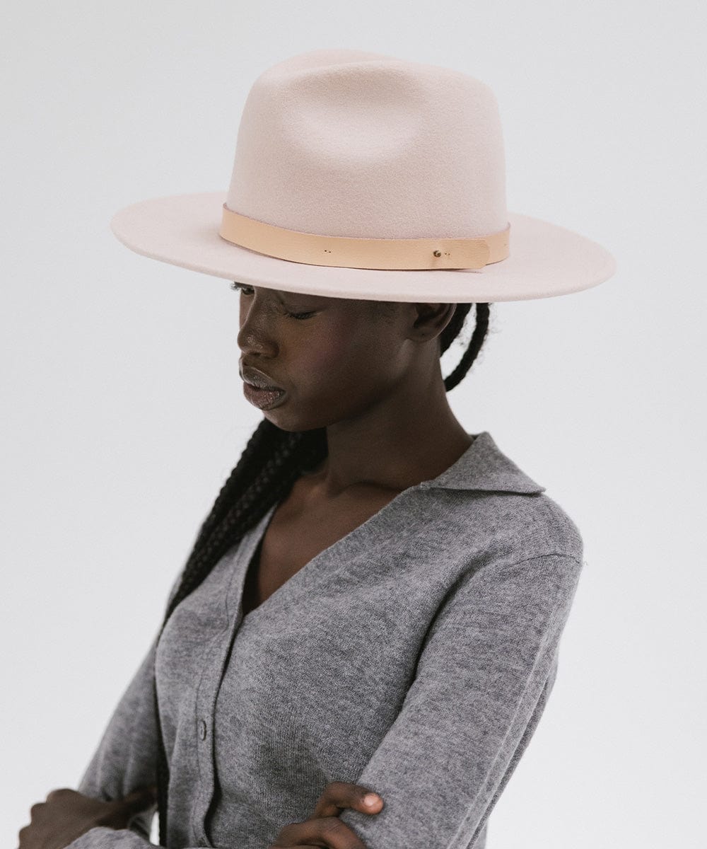 Woman wearing a blush pink colored fedora hat with a hat band and a grey shirt, looking sideways against a plain background #color_blush pink