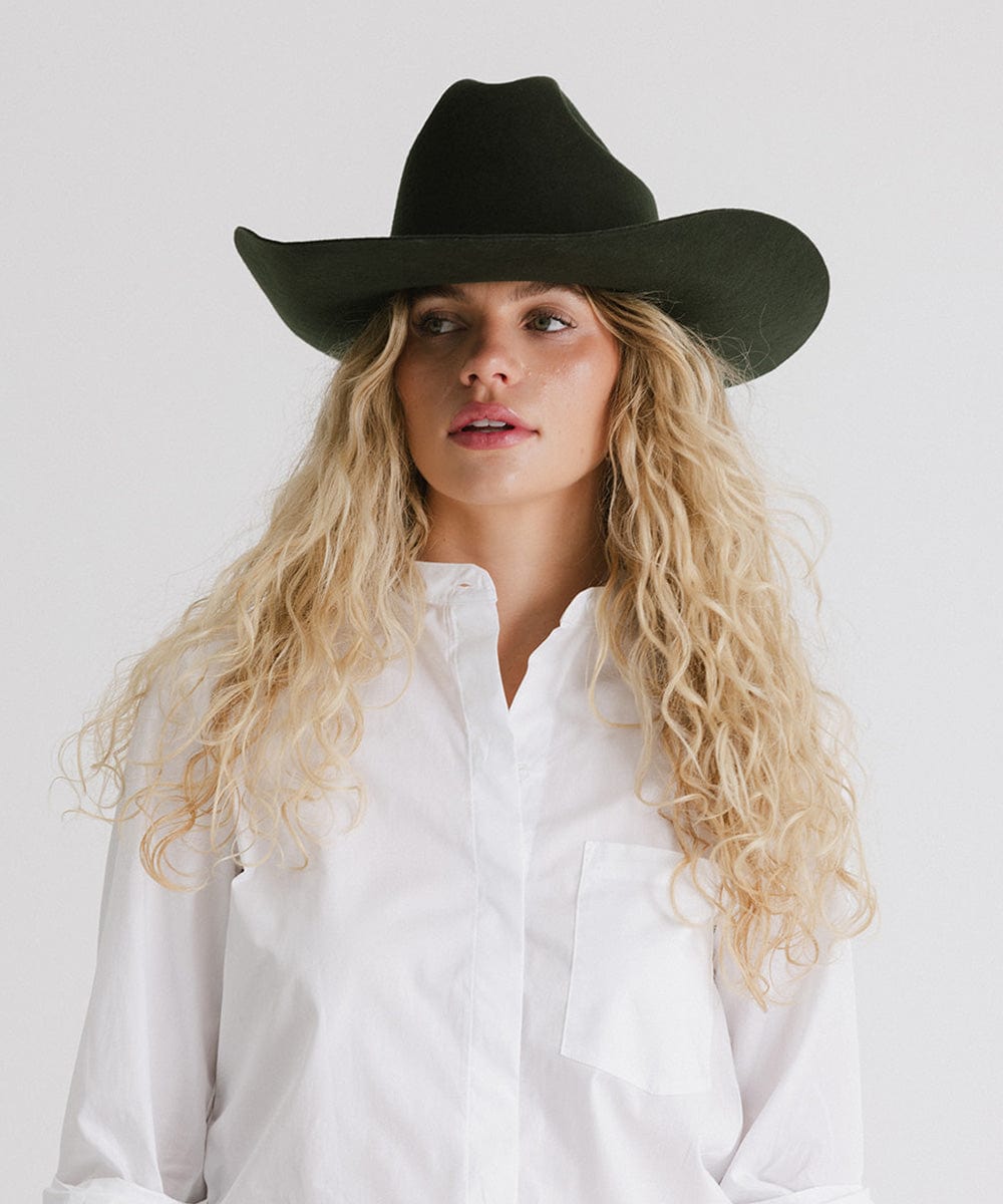 Woman wearing a dark green cowboy hat and a white shirt against a plain background #color_dark green