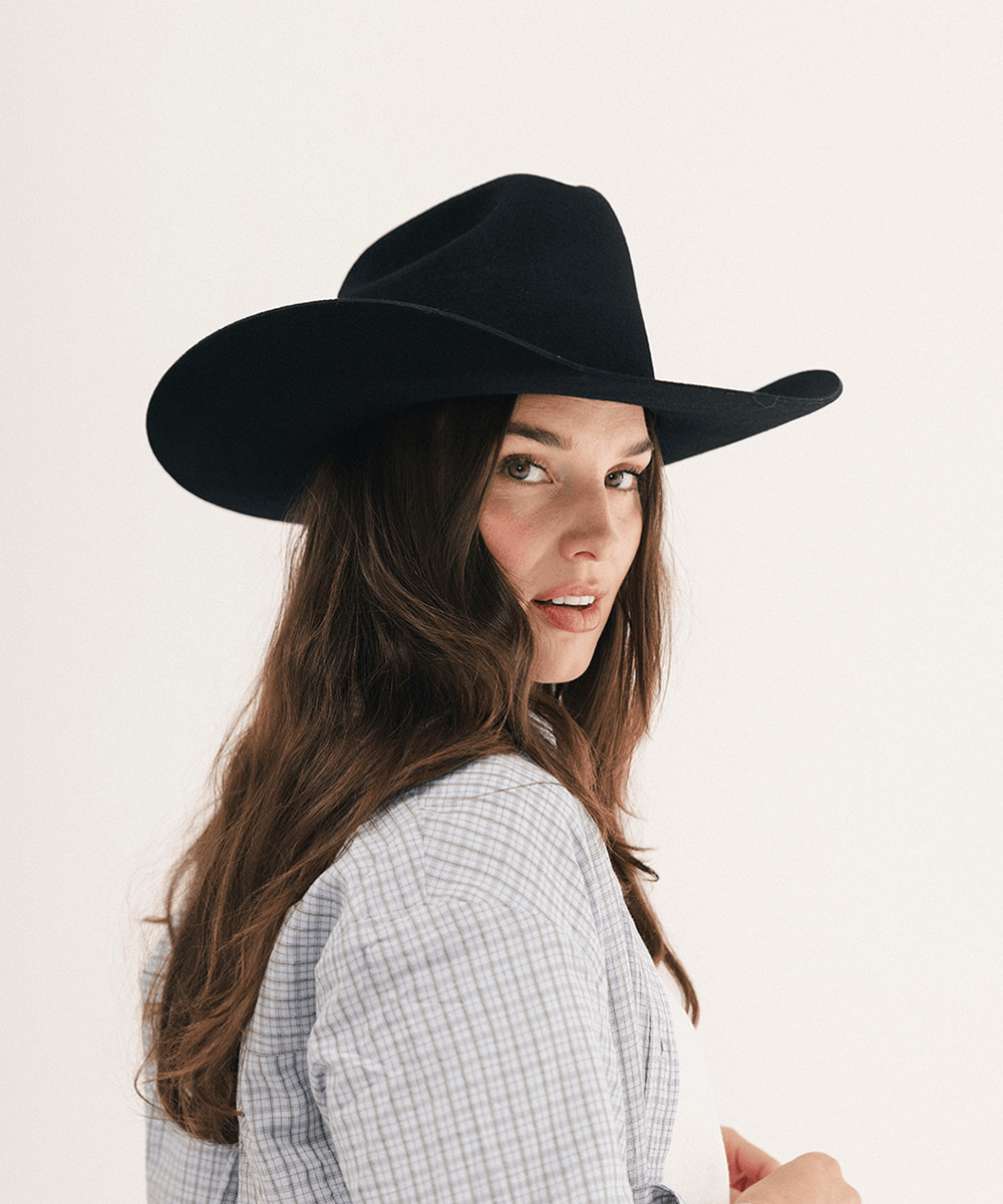 A woman wearing a black felt cowboy hat and a light blue and white checkered shirt, photographed against a clean off-white background #color_black