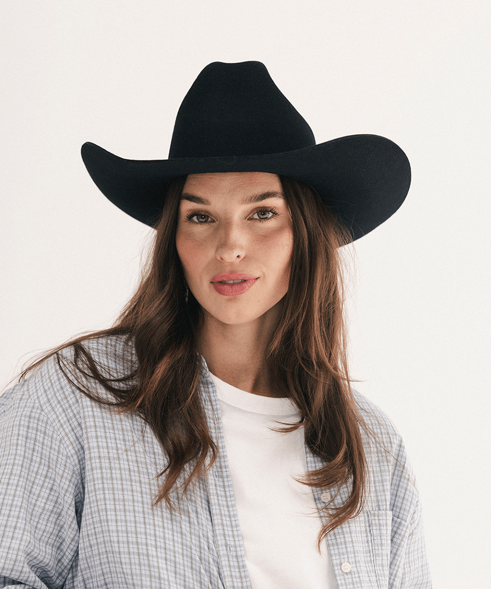 A woman wearing a black cowboy hat, a white t-shirt, and a light blue and white checkered button-up shirt, photographed against a clean white background #color_black