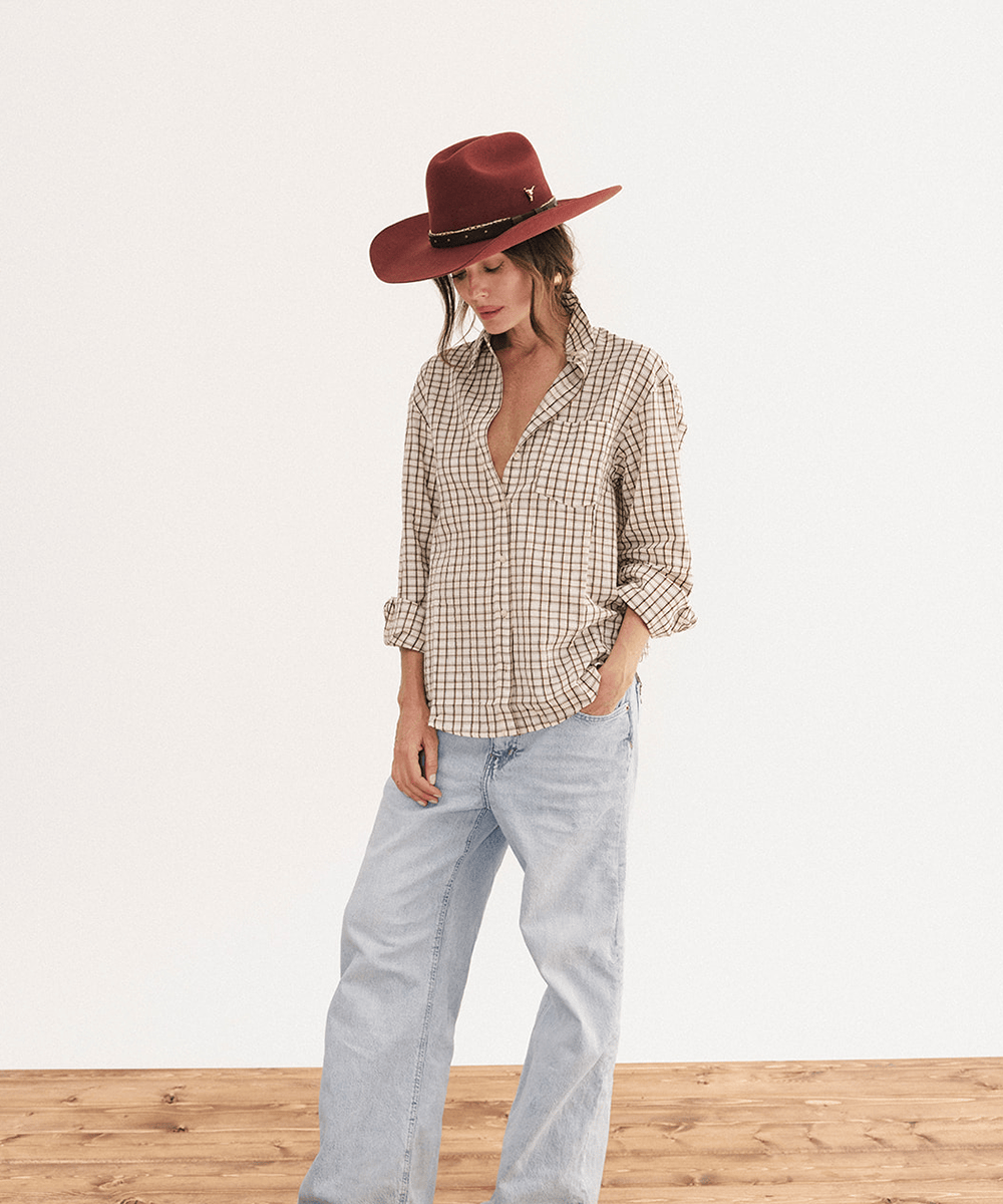 A woman stands in a studio wearing a brown and beige gingham button-up shirt, light-wash wide-leg jeans, and a burgundy wide-brim hat against a white wall and wood floor #color_rusty red