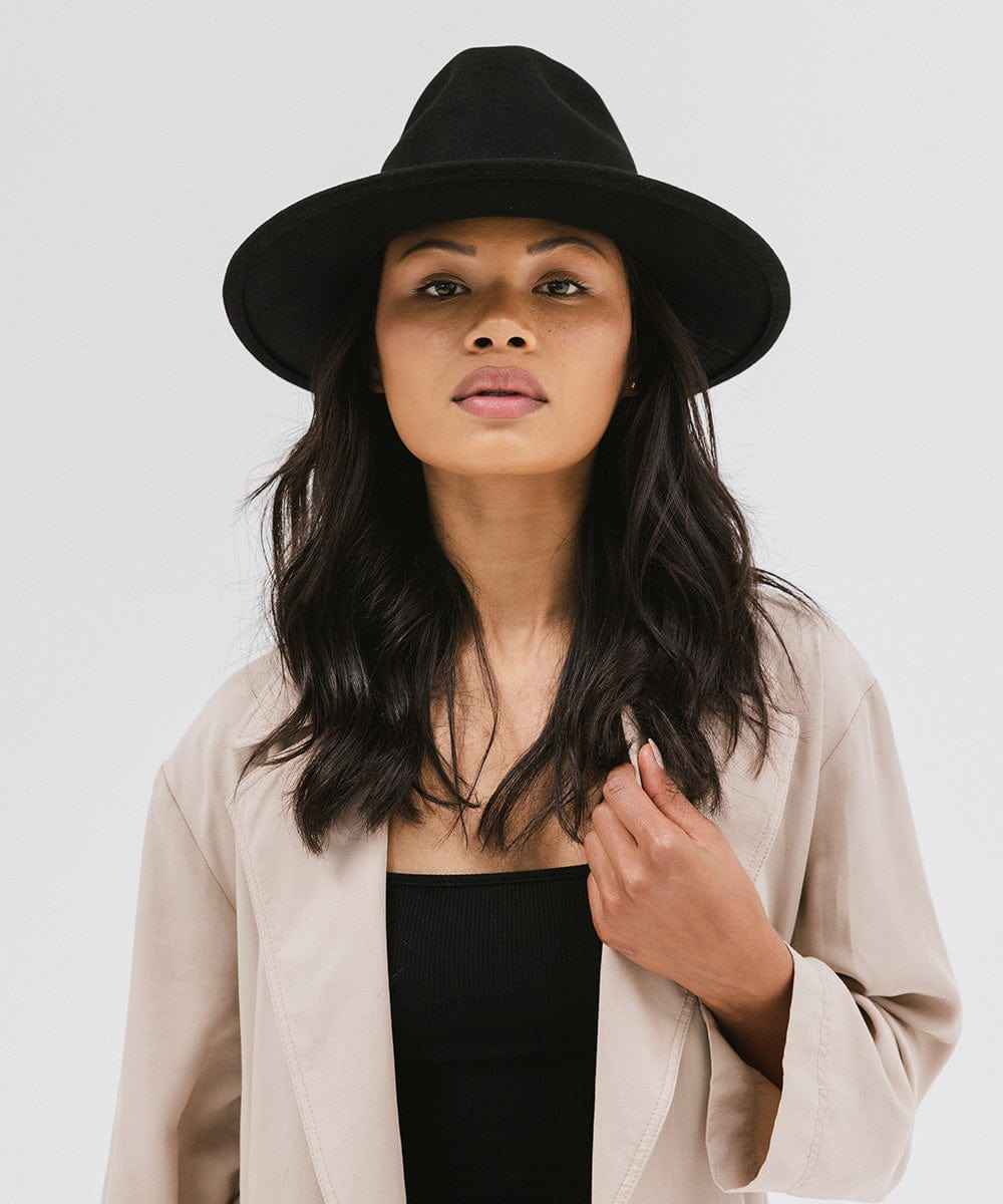 Woman wearing a black fedora hat and a beige coat with a black top against a plain background #color_black