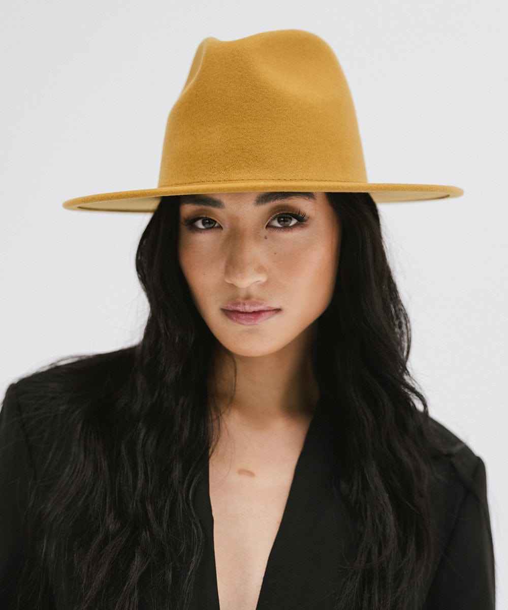 Woman wearing a mustard colored fedora hat and a black coat against a plain background #color_mustard
