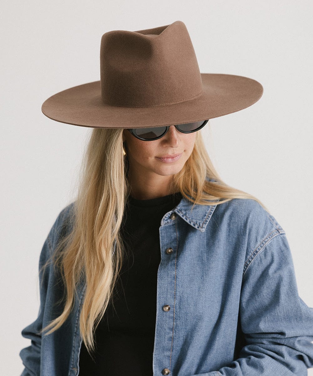 Woman wearing a chocolate colored fedora hat, sunglasses and a black t-shirt with a denim shirt, looking sideways against a plain background #color_chocolate
