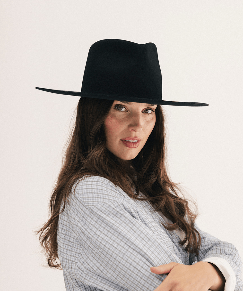 A woman wearing a black wide-brim fedora hat and a light blue and white checkered shirt, photographed against a plain off-white background #color_black