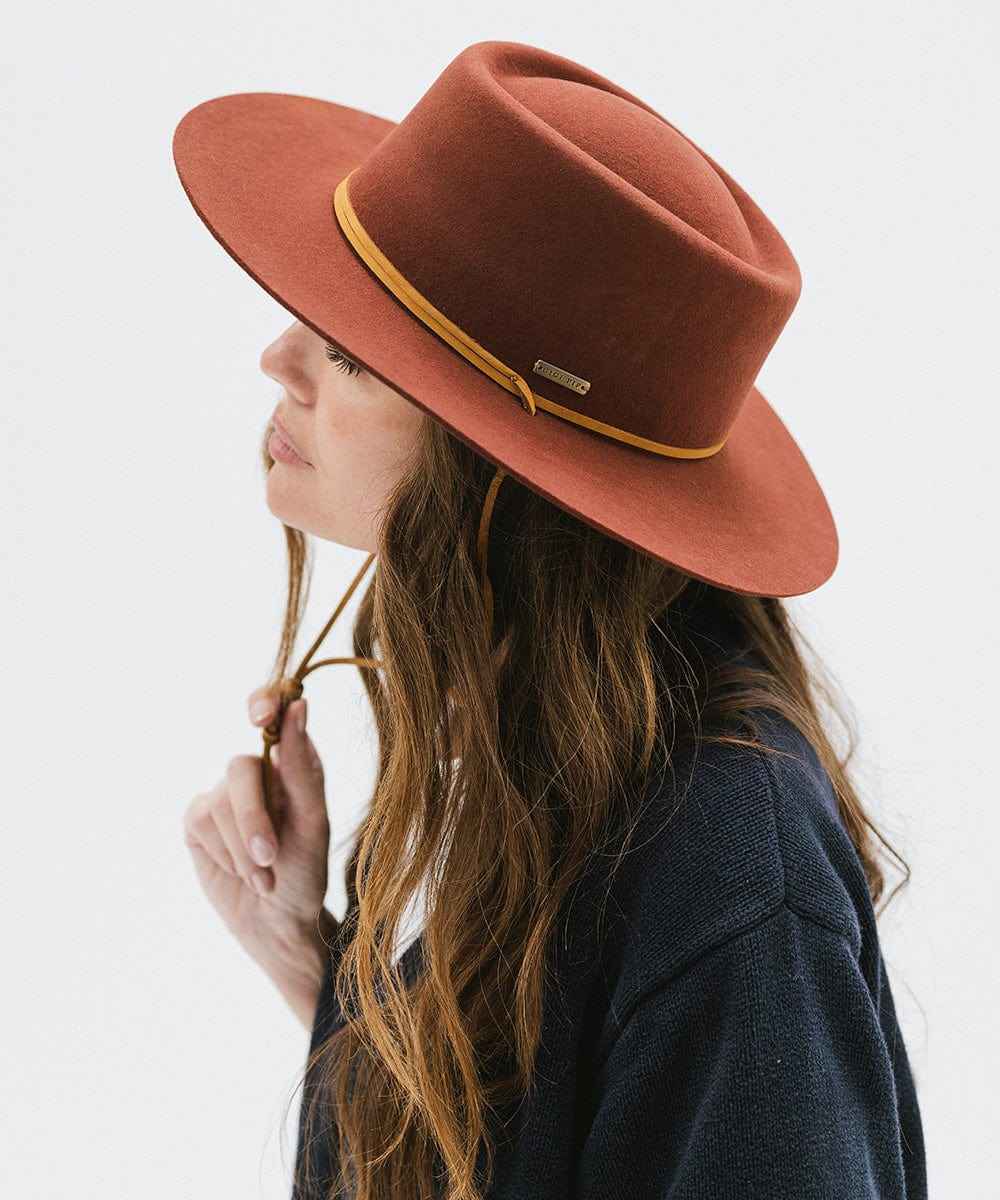 Woman wearing a rusty red colored fedora hat with a yellow band, facing sideways against a white background #color_rusty red