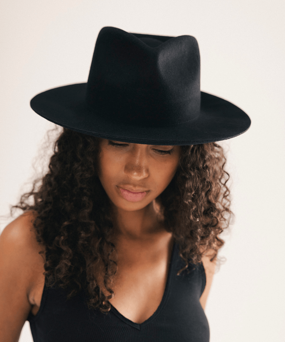 A woman with long curly brown hair wearing a black wide-brim fedora hat and a black tank top, photographed against a plain light background #color_black