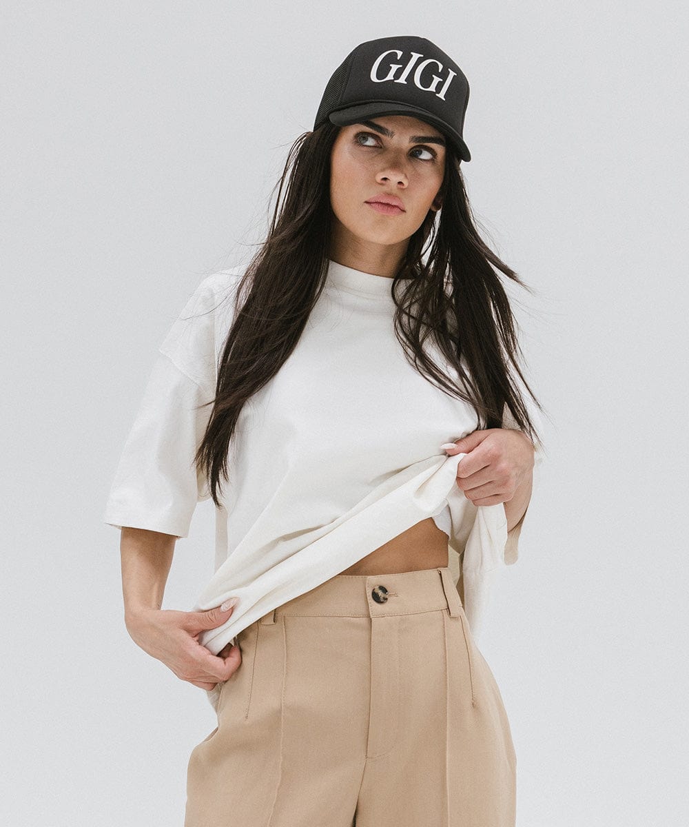 Woman wearing a black cap with 'GIGI' text, a white t-shirt and beige pants against a plain background #color_black