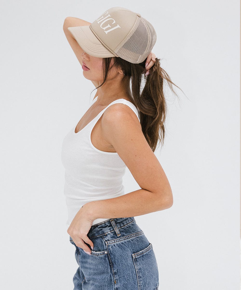 Woman wearing a tan cap with 'GIGI' text, a white top and blue jeans, posing sideways against a plain background #color_tan