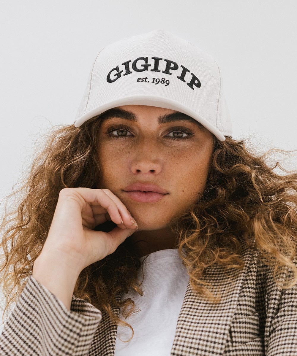Woman wearing a cream trucker hat with 'GIGI PIP' embroidered in black on the front, and a white top with a coat against a plain background #color_cream