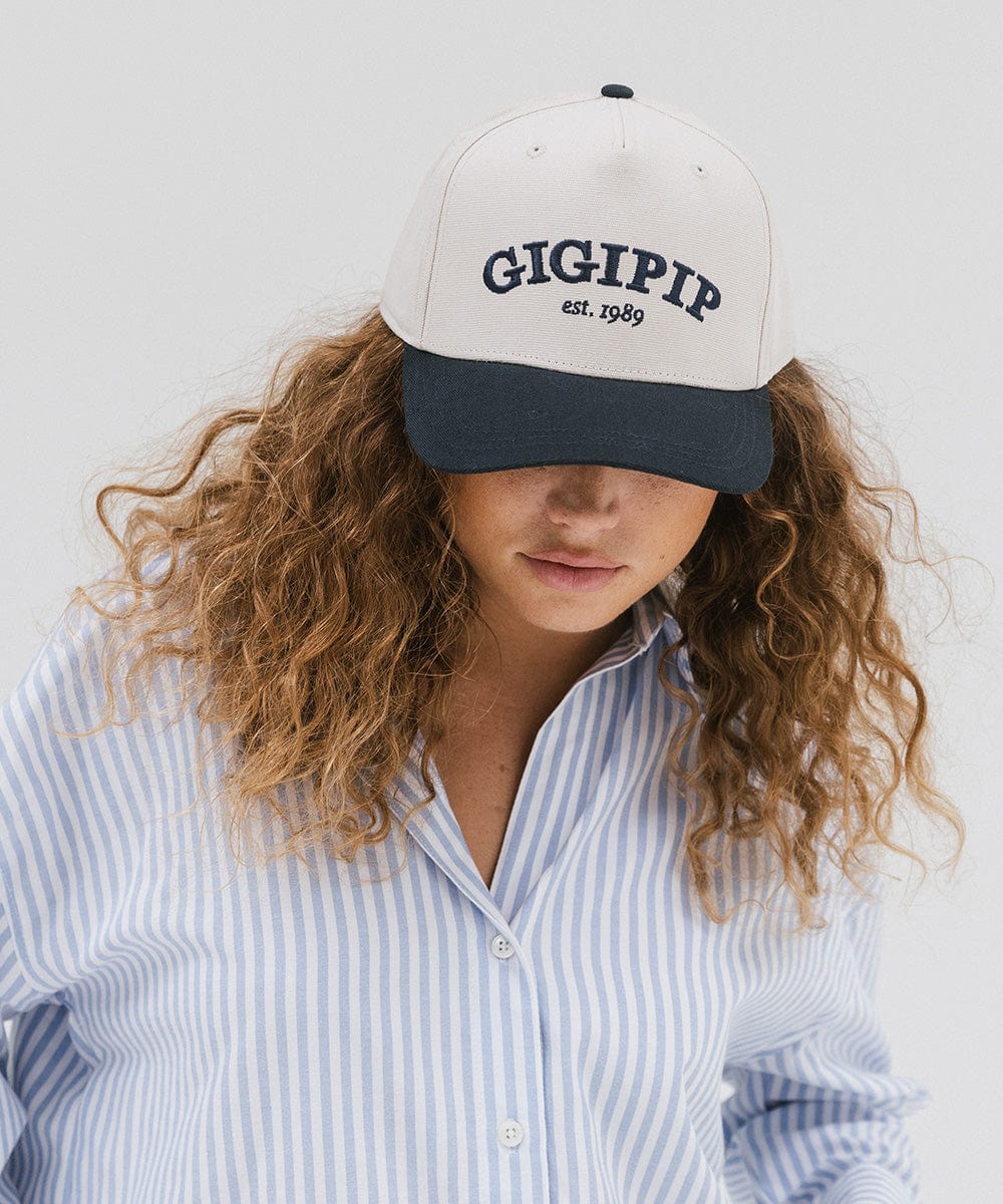 Woman wearing a cream and navy trucker hat with 'GIGI PIP' embroidered in navy on the front and a light colored shirt against a plain background #color_cream - navy