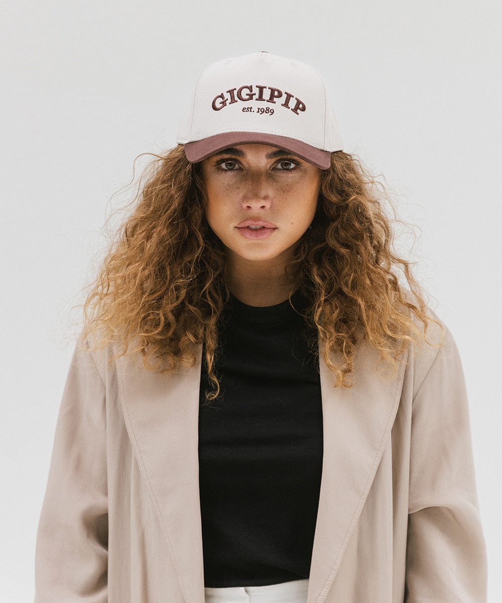 Woman wearing a cream and chocolate brown trucker hat with 'GIGI PIP' embroidered in chocolate brown on the front and a black top with a beige coat against a plain background #color_cream - chocolate brown