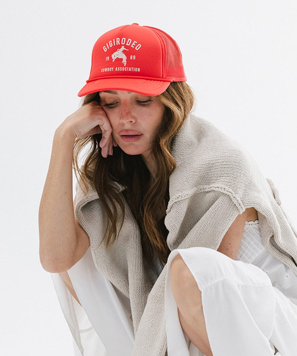 Woman wearing a vintage red cap with golden 'GIGIRODEO' text and logo, and a grey sweater and white pants, sitting against a plain background #color_vintage red
