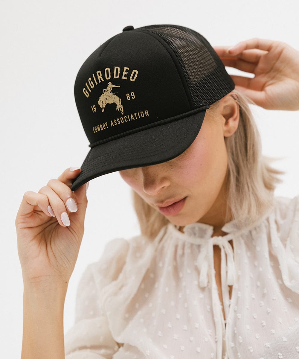 Woman wearing a black cap with golden 'GIGIRODEO' text and logo, and a white top against a plain background #color_black